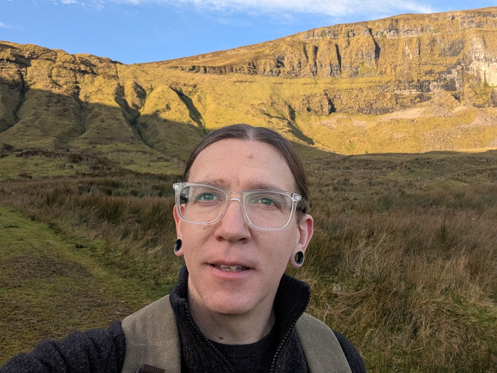An image of Meadow, owner and creator of Rainbow Crow Books & Games, who has long brown hair parted in the middle and pulled back into a bun. They wear clear glasses and have black open ear gagues. They are standing in front of a mountain bluff in Ireland. They are smiling and almost about to speak in the photo.